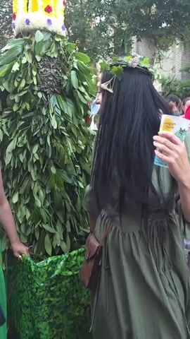 The video shows a person dressed in a costume made entirely of green leaves, resembling a large tree or bush. The costume is cone-shaped and adorned with a large, elaborate crown at the top, which is yellow and decorated with colorful flowers or fabric. Two women are visible in the foreground, interacting with the leaf-costumed figure. The woman on the right has long, dark hair and wears a green dress and a floral wreath on her head. The woman on the left has curly hair and is wearing a green dress and a white top. She holds a dark handbag. Both women appear to be holding onto the lower part of the leaf costume. In the background, other people can be seen, some in festive attire, and there are trees and buildings suggesting an outdoor gathering or festival. The overall atmosphere seems lively and celebratory.