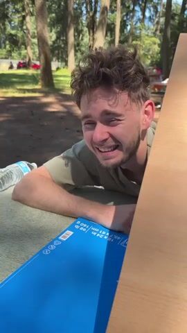 A young man with brown curly hair lies on his stomach on a light green mat, with his head resting on his folded arms. He is outdoors in a wooded area with sunlight filtering through the trees. In the foreground, a blue cardboard box with text printed on it lies on the mat to the left of the man. A wooden board is positioned to the right, obscuring part of the man's face. A plastic water bottle is visible on the mat behind the man's head. In the background, several people are seated on chairs or lying on blankets in the grassy area under the trees. A red car is parked in the far background to the left. The man opens his mouth wide and lets out a loud scream. He then lowers his head onto his arms and appears to fall asleep.