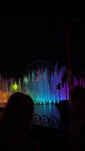 The video shows a night scene with a water fountain show featuring colorful lights. Water jets of various sizes and shapes are illuminated in a rainbow of colors, including blue, green, purple, red, and yellow. A Ferris wheel is visible in the background, also lit up with colorful lights. Silhouetted figures of people are visible in the foreground, suggesting they are watching the show. A decorative railing runs along the edge of the water, separating the viewers from the display.