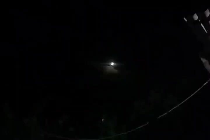 The video displays a nighttime scene. In the foreground, a hand forms a small heart shape with its thumb and index finger. Beyond the hand, the moon is visible as a bright, glowing orb in a dark sky. Silhouettes of foliage are present in the background, and the overall lighting is dim, illuminated by the moon.
