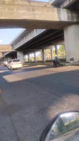 A daytime, eye-level, outdoor, medium shot captures traffic flowing beneath an elevated concrete overpass. The road is asphalt with visible lane markings. On the right side of the frame, a person in a green shirt and helmet rides a light-colored scooter away from the camera. Further down the road, another scooter rider in a red helmet and white shirt heads towards the camera. In the background, several other vehicles, including cars and scooters, are visible. The overpass, supported by concrete pillars, casts a shadow on the road. The sky is clear and blue. A wooden table with items on it sits on the left side of the frame in the foreground.