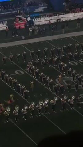 The video shows a marching band on a football field. The band members are wearing uniforms and playing instruments. They are marching in formation. The video is taken from an overhead perspective. The lighting is bright. There are people in the stands in the background.