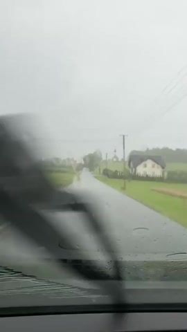 The video shows a view from inside a vehicle driving on a wet, paved road. The sky is overcast and appears to be raining, with water droplets visible on the windshield. The road is lined with green fields and some houses and trees in the distance. There are also power lines visible above the road. The vehicle appears to be moving forward at a moderate pace.