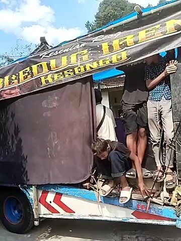 The video shows a group of people in what appears to be a rural setting. In the first few seconds, a truck with a black tarp covering its cargo area is visible, with several young men standing on and around it. The truck has yellow lettering on its tarp, and some of the men are wearing casual clothing. The scene then shifts to a courtyard in front of a building with a light-colored roof and a green awning. A group of people, many of whom are wearing traditional costumes with colorful fabrics and headdresses, are gathered in the courtyard. Some individuals are holding what appear to be musical instruments or props. The overall atmosphere suggests a cultural performance or event is about to take place or is in progress. The ground is dirt and debris is scattered around. The background shows trees and houses, indicating a village or countryside location.