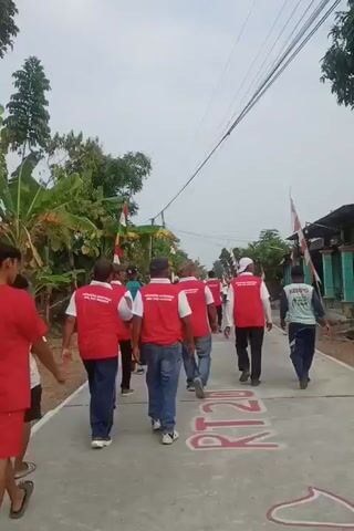 A group of people are walking down a paved road. Most of the individuals are wearing red vests with white lettering, suggesting they are part of an organization or team. The vests have text printed on them. Some individuals are wearing blue jeans, and others are wearing shorts. The road has markings painted on it in red, with the letters "RT" and numbers visible. There are trees and vegetation on the sides of the road, and houses or buildings can be seen in the background. Several Indonesian flags are also visible, attached to poles and structures. The sky is overcast.