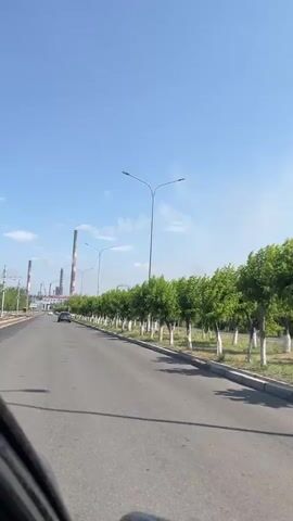 The video shows a view from the inside of a car traveling down a paved road. The road is lined with trees on the right side, and there are streetlights along the road. In the distance, several tall industrial smokestacks are visible against a clear blue sky with a few white clouds. A car is also visible on the road in front of the camera car. The overall environment appears to be an industrial area with some greenery.