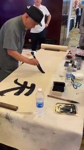 A man in a grey traditional robe and a black hat is practicing calligraphy on a long scroll of paper. He is holding a brush with black ink and making strokes on the paper. There are several bottles of water, ink wells, brushes, and a pair of scissors on the table. In the background, there are other people, one of whom is taking a picture of the calligrapher with his phone. The room appears to be a well-lit indoor space, possibly a conference room or an exhibition hall. The overall atmosphere seems to be one of concentration and artistry.