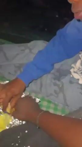 A young man in a blue sweatshirt sits cross-legged on a blanket, eating from a foil package. He is sitting close to another person, presumably a friend, who shares the package. They are outdoors, likely at night, as the light is dim. The young man is the focal point of the video, and he looks directly at the camera at one point. The foil package appears to contain food wrapped in something yellow.