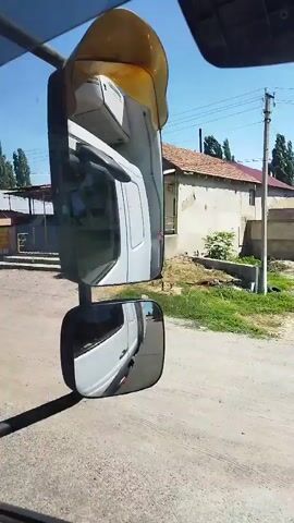 The video begins with a view from inside a vehicle, looking at the side mirrors. The right side mirror shows a white truck cab reflected. The left side mirror shows a white truck cab and a house with a beige facade. The camera then shifts to the inside of the truck, looking through the windshield. The truck is driving on a paved road. On the road ahead, a truck is transporting a large stack of hay bales. To the left of the hay truck, a black car is driving. To the left of the black car, another truck, this one smaller and white, is driving. The road is lined with trees on both sides. The sky is blue and clear. The video ends with a continued view of the truck with the hay bales driving on the road.