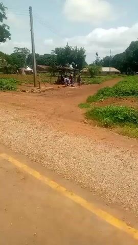 The video shows a dusty, rural road with a yellow line painted down the center. On one side of the road, there is a small cluster of buildings, some appearing to be made of mud brick with corrugated metal roofs, surrounded by green vegetation and trees. A few people are visible near one of the structures, seemingly engaged in some activity. The other side of the road is largely undeveloped, with some patches of dirt and low-lying greenery, leading towards more trees and open space under a cloudy sky. The camera appears to be mounted on a moving vehicle, as the perspective shifts and the background blurs slightly.