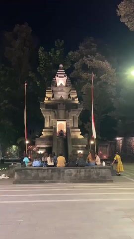 A group of approximately ten people are seated on a tiered stone platform at night, facing a large, illuminated stone structure. The structure resembles a temple or monument with multiple tiers and intricate carvings, topped with a spire. Two flags, one red and white and the other mostly white with a red stripe, are positioned on flagpoles on either side of the structure. Small lights are visible around the base of the monument. Trees are visible in the background, and the sky is dark, indicating nighttime. One person, wearing a yellow dress, is standing on the right side of the frame, observing the monument. The overall scene appears to be a public gathering or a moment of contemplation in front of a significant landmark.