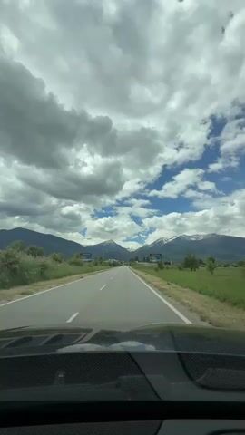 The video is shot from inside a car, looking out through the windshield at a highway. The road is asphalt and has white lane markings. On either side of the road are grassy fields and some trees. In the distance, there are mountains, some of which have snow on their peaks. The sky is partly cloudy with patches of blue sky visible. There are two billboards visible on the sides of the road. The car appears to be moving.