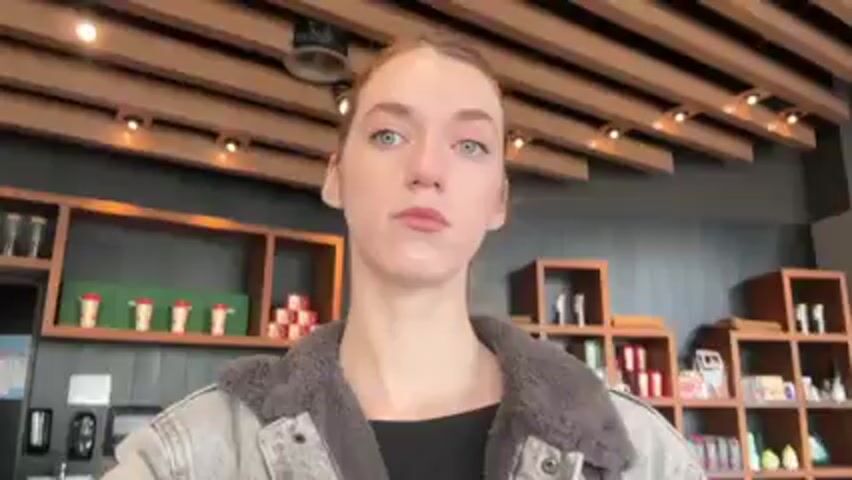 The video shows a person with fair skin and light blue eyes, wearing a black top and a light grey jacket with a dark grey fur-like collar. The person is standing indoors, likely in a commercial establishment. In the background, there are wooden shelves stocked with various products, including cups, boxes, and bottles. To the left, there is a refrigerator and what appears to be a point-of-sale system. The person looks directly at the camera with a neutral expression. The lighting is bright, and the scene appears to be well-lit.