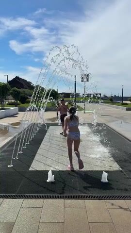 The video captures an outdoor scene on a bright, sunny day, featuring a ground-level water fountain. A clear blue sky with scattered white clouds is visible overhead.

In the center of the frame, two children and an adult are playing in the water fountain. A young girl, wearing a patterned two-piece swimsuit, runs through the arching water jets, appearing to be enjoying herself. A young boy, dressed in dark swim shorts, is also seen running and splashing in the fountain. An adult male, similarly in dark swim shorts, stands within the fountain, interacting with the children.

The fountain consists of multiple water jets emerging from the paved ground. There's a prominent arch of water created by several jets on one side, and individual upright geysers of water elsewhere. The ground around and within the fountain is paved with light-colored tiles and dark metal grates.

Beyond the fountain area, there are green grassy sections, some small trees, and paved walkways, suggesting a park or public square. In the distance, hills or buildings can be observed under the bright sky. The overall atmosphere is lively and joyful, with the individuals engaging in recreational water play. No discernible text is visible in the video.