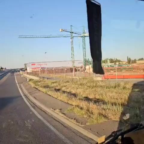 The video shows a view from a vehicle moving on a road, driving past a large building identified as "Premier Pigs." The building is white and gray with a prominent red logo of a pig silhouette and the company name in red letters. A metal fence surrounds the property, and a "100" speed limit sign is visible. In the background, there are construction cranes and other building materials, suggesting an ongoing construction project nearby. The sky is clear and blue.