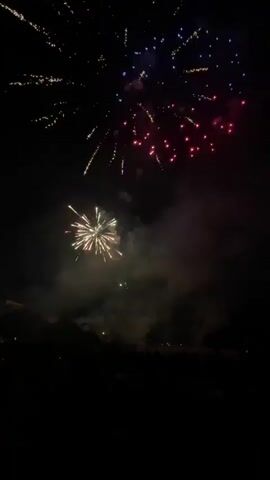 The video shows a fireworks display at night. The sky is dark, illuminated by bursts of colorful fireworks. In the foreground, a crowd of people is silhouetted against the bright explosions. The fireworks are primarily red and gold, with some green and pink accents. The smoke from the explosions hangs in the air, creating a hazy atmosphere. The camera is positioned at ground level, looking up at the fireworks. The video is short, lasting only a few seconds.