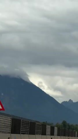 The video shows a view from a vehicle on a highway, looking out at a mountain range under a cloudy sky. A sound barrier runs along the side of the highway. Cars and trucks are visible on the highway, moving in both directions. The mountains are dark blue and green, with some clouds obscuring the peaks. The sky is overcast with gray clouds. There are trees and other vegetation visible on the slopes of the mountains and along the side of the highway. The overall impression is one of a scenic drive on a cloudy day.