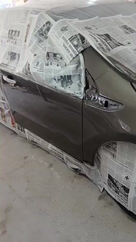 The video pans across the front portion of a car that is being prepped for painting. Newspaper is used to mask off various parts of the car, including the headlights, front wheel wells, and the hood. The car appears to be a dark gray color, and the newspaper is taped securely in place. The environment appears to be a workshop or garage, with tools and equipment visible in the background.