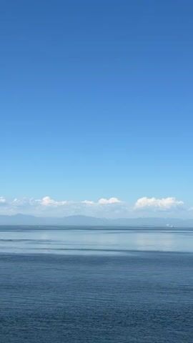 The video shows a serene seascape under a clear blue sky. The horizon is marked by a thin strip of distant land with mountains, partially obscured by a hazy atmosphere. Below the landmass, a calm, deep blue ocean stretches out, reflecting the light and showing subtle ripples on its surface. A few fluffy white clouds are scattered along the horizon, adding a touch of texture to the vast expanse of the sky. The overall impression is one of tranquility and natural beauty.