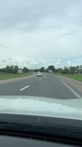 A dash cam perspective shows a gray SUV driving on a paved road. The road curves to the right and is bordered by a grassy shoulder on both sides. There are houses and trees visible in the background. Several other vehicles are on the road, including a white Volkswagen, a gray Peugeot, and two other cars further in the distance. The sky is cloudy.
