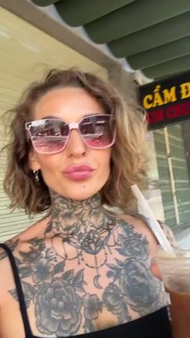 A woman with extensive tattoos on her chest and neck, wearing a black tank top and pink sunglasses, holds up a plastic cup with a brown beverage and a straw to the camera. She smiles with her lips slightly parted, revealing her teeth. Her short, wavy, light brown hair is visible. The scene appears to be outdoors, with a building facade and a sign in Vietnamese visible in the background. The lighting suggests it is daytime. The overall mood is casual and cheerful. The visible text on the sign appears to be "CẨM ĐỒ" and "TIN CHUNG".