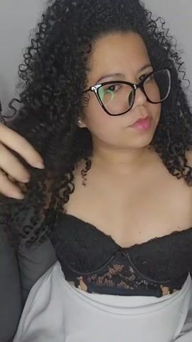The video features a woman with curly dark hair and black glasses. She is wearing a black lace top and a white garment covering her lap. The background appears to be a gray couch and a white wall. She is looking at the camera and holding her hair with one hand. She purses her lips, possibly blowing a kiss.