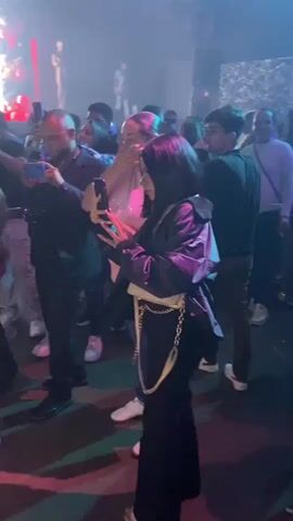 The video shows a busy and crowded event, possibly a party or a concert. There are people dressed in various styles, many holding phones and recording the event. In several shots, people are taking photos, and a red carpet entrance with balloons suggests a formal event. People are smiling and appear to be enjoying themselves, creating a lively atmosphere.