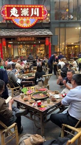 The video presents a bustling outdoor dining area of a Chinese restaurant called 走起川 (zou qi chuan).  Several groups of people are seated at wooden tables with bamboo chairs, enjoying a meal. Many of the tables hold large dishes of food, including what appears to be a spicy hot pot with red broth, as well as smaller bowls and tea sets. People are seen eating, drinking, talking, and using their phones. Waitstaff in blue uniforms are moving between tables, serving customers. The restaurant has a traditional Chinese architectural style with red lanterns and decorative signage.  The dining area is surrounded by modern buildings with glass windows, creating a contrast between the traditional restaurant setting and the modern cityscape.