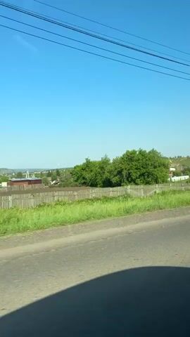 The video shows a view from inside a moving vehicle, looking out at a rural landscape. The time of day is daytime, with a clear blue sky. The foreground consists of a paved road with a shadow of the vehicle on it. Beyond the road, there is a grassy embankment and a wooden fence. In the middle ground, there are several houses with various roof colors, some surrounded by trees. There is a prominent utility pole with power lines. In the background, there are rolling hills. A silver car is visible driving on the road in the middle distance. The overall impression is a sunny day in a village or countryside setting.