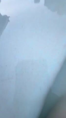 The video shows a person's legs submerged in a swimming pool. The water is clear and blue. The person's legs are crossed at the knees. The camera appears to be held underwater, looking up at the person's legs. In the background, there is a pool deck, a hedge, and some pool equipment. The video is short, lasting only a few seconds.