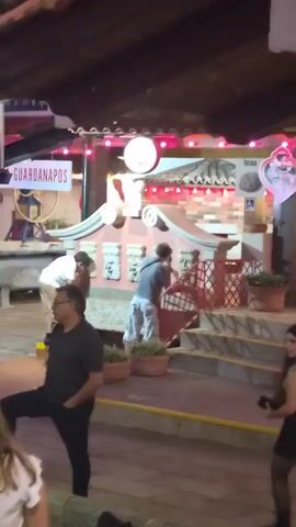 The video shows a group of people in what appears to be an outdoor dining or entertainment area at night. Colorful string lights are visible overhead. In the background, a sign reads "GUARDANAPOS." There are decorative architectural elements on a pink wall, including what look like carved faces with spouts. One person in a white shirt and tan pants is bent over, appearing to drink from one of the spouts. Several other individuals are visible, some standing and some walking, in the foreground and mid-ground. The ground is wooden planking.