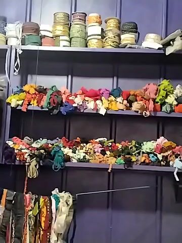 The video displays an interior shot of a storage area or workshop, likely for textiles or crafts. Three prominent shelves are visible against a dark purple background. The top shelf holds numerous spools of thread or ribbon in various colors, including white, gold, green, red, and brown. A string of white beads hangs on the left side of this shelf, and a few white caps or hats are placed on the right. The middle and bottom shelves are filled with large, disorganized piles of colorful fabric scraps or remnants, featuring a wide spectrum of colors such as red, pink, yellow, green, blue, orange, and brown. At the very bottom of the frame, several pieces of clothing or fabric are seen hanging on a rod. The shot is static, with no people or activity present. There is no visible text in the scene.