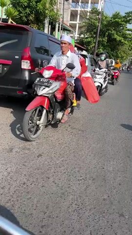 The video shows a street scene with several vehicles and people. In the foreground, a black SUV is partially visible on the left. A white scooter with a rider wearing a light brown hijab and a face mask is in the center of the frame. To the right of the white scooter, a red scooter with a rider wearing a yellow hijab and a dark green outfit is also present. Further back, a man on a black scooter is visible. The street appears to be paved with asphalt, and there are buildings and trees in the background. The overall impression is a moderately busy street, possibly during daytime given the lighting.