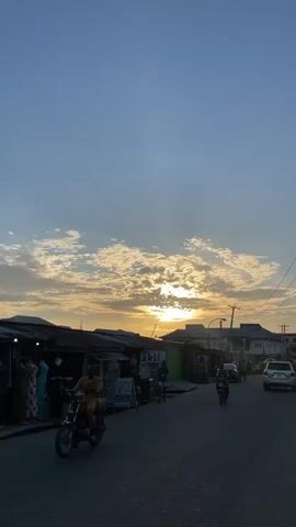 The video shows a street scene at sunset. The sky is blue with scattered white clouds, and the sun is visible behind the clouds, creating a golden glow. On the left side of the street, there are small shops with corrugated iron roofs. People are standing in front of some of the shops, while others walk by. On the right side of the street, there are cars parked and a motorcycle drives by. In the background, there are taller buildings. The road is paved and appears to be in a busy urban area.
