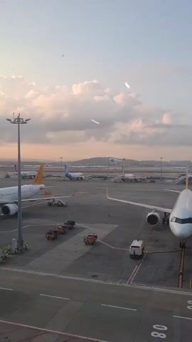 The video shows an airport tarmac scene captured from an elevated viewpoint, possibly from inside an airplane or a building overlooking the tarmac. The time of day appears to be either sunrise or sunset, indicated by the warm, orange glow of the sky and the soft light illuminating the clouds. Several airplanes are visible on the tarmac, including a prominent white and orange Pegasus Airlines plane parked in the foreground, with a jet bridge attached. Other aircraft, including a blue and white airplane and a plane with a Turkish Airlines logo, are parked or moving in the background. Several ground support vehicles, such as baggage carts and a utility vehicle, are also present. In the distance, an airplane is ascending into the sky, leaving a contrail. The overall environment is an active airport tarmac under a cloudy, atmospheric sky.