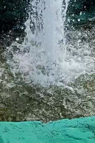 The video shows a fountain with water spraying upwards. The fountain appears to be made of rock-like structures covered in green moss. The water is clear and turbulent, creating a spray of droplets. The background is a mix of blue sky with white clouds and some greenery. There are no people visible in the video.