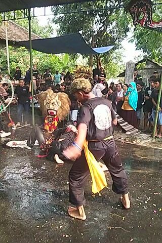 The video shows a man in black clothing with a yellow sash performing a martial arts move over another person dressed in black who is lying on the ground. The man is barefoot and has curly hair. The performance is taking place on a wet, paved surface with a crowd of people watching in the background. There is a decorative object to the left of the frame and trees are visible in the background.