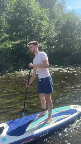A young man with short brown hair and a beard stands on a paddleboard in a river. He is wearing a white t-shirt and blue shorts. He holds a paddle in his right hand, and his left hand is near his chest. The paddleboard is blue, white, and teal. There is a black bag on the front of the paddleboard. The river has visible ripples and submerged vegetation. Lush green trees line the riverbank in the background. The sky is visible above the trees. The man appears to be moving slowly through the water.