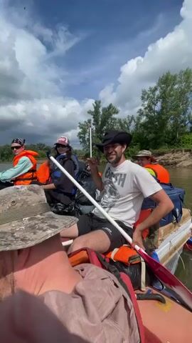 A group of five people are on a raft on a body of water. The sky is partly cloudy and the sun is shining. The people are wearing life vests and casual clothing. One person in the foreground is wearing a camouflage hat and sunglasses. Another person is wearing a cowboy hat and a white t-shirt with a graphic. A third person is wearing a purple baseball cap. The raft is red and white. There are paddles in the water. The scenery appears to be a riverbank with trees. The person in the foreground is holding up a bottle.