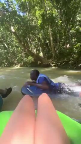 A first-person perspective video shows two people tubing down a river in a lush, green environment. A man in a blue and purple shirt and a life vest is in the water, holding onto the front of another blue tube that is holding a person with fair skin and colorful shorts. The person filming is in a green tube. The river is flowing, and the trees create a canopy overhead. The man in the water appears to be guiding or assisting the tubes as they move down the river. The person recording is floating downstream.