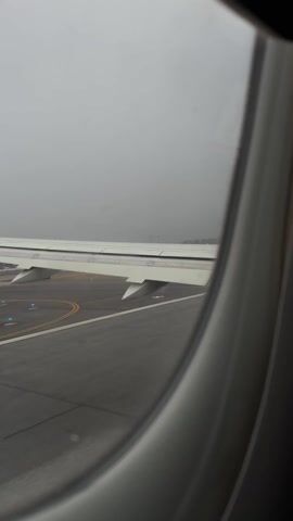 The video is a view from inside an airplane, looking out the window. We see the plane's wing and flaps, as well as the airport runway and surrounding area. The weather is overcast and visibility is limited. The runway markings are visible, and in the distance, we can make out other airport buildings and possibly other airplanes. The airport control tower is visible in the distance. The plane is stationary.