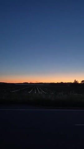 The video shows a dark, rural landscape at twilight. A black asphalt road with a white stripe runs horizontally across the bottom of the frame. Beyond the road is a field, and beyond that, a silhouette of trees against a deep blue and orange gradient sky at dusk. As the camera pans to the right, more trees are visible, and a road sign is seen on the right side of the road.