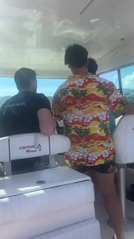 Two men are on a boat. One man, wearing a black t-shirt with "Calvin Klein" printed on the back, sits at the helm, looking out a window. The other man, wearing a colorful Hawaiian shirt and patterned shorts, stands beside him, facing away from the camera. The boat's interior is white and appears to be clean. Sunlight streams in through the windows, indicating it is daytime and likely warm. The water is visible through the windows, suggesting the boat is in motion.
