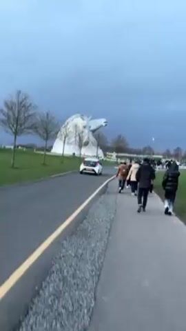 The video depicts a view of The Kelpies sculptures in Scotland under a dark, overcast sky. A small group of people is walking along a paved path next to a road with a white car driving on it. The Kelpies, two large horse-head sculptures, are visible in the background across a grassy field. Bare trees line the field. The overall atmosphere is somewhat bleak due to the weather.