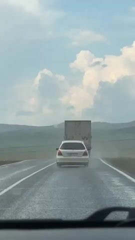 This is a first-person perspective video taken from inside a vehicle driving on a wet highway. The viewpoint is looking through the windshield, which has a windshield wiper visible in the foreground. The road is wet and appears to be raining, with water splashing up from the tires of vehicles. In front of the camera vehicle, a white car is visible, also driving on the highway. Ahead of the white car, a large truck with a trailer is driving in the same direction. The truck's trailer is light-colored and appears to be made of a material like canvas or plastic. The truck and car are both creating spray as they drive through the water on the road. The environment appears to be rural, with rolling hills and green fields visible in the background. The sky is cloudy with patches of blue sky visible. The video is shot in landscape orientation. The timestamp indicates the video is very short, only a few seconds long.