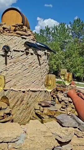 This is a vertical video of a decorative outdoor fountain designed to resemble a wine theme. The fountain is constructed with stacked stones and rocks. At the top, a wooden barrel is visible, and two dark-colored wine bottles are positioned to appear as though they are pouring liquid. The liquid, which appears to be light-colored, possibly wine or water, flows down into large, clear wine glasses that are also part of the fountain's design. The glasses are positioned on different levels of the stone structure, and the liquid collects in them. In the foreground, a large, reddish-brown clay amphora is placed among the rocks. The background shows green trees and a bright blue sky with some white clouds, indicating an outdoor setting on a sunny day.
