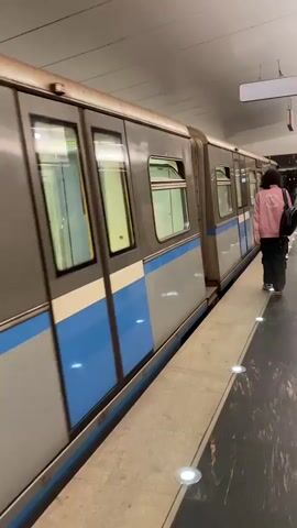 The video captures a scene on a well-lit subway platform in what appears to be a Russian subway station. The platform features light-colored floor tiles with embedded circular lights and a darker, possibly marble, edge alongside the tracks. The ceiling is white and illuminated by fluorescent lights.

Initially, a grey and blue subway train is stationary at the platform. The train's doors are closed, and a distinct, loud mechanical sound suggests it is preparing for departure. A person with long, dark hair, dressed in a pink or light red jacket, dark pants, and carrying a black backpack, walks along the platform beside the train, moving away from the camera.

The camera then shifts focus to a man walking in the same direction. He is wearing a red short-sleeved shirt with a "Russell" logo, blue jeans, and a black backpack. He holds a dark cup in his right hand. The train remains visible on the left, and the number "75" can be seen on its side. Several other individuals are also walking further down the platform in the distance. The setting is indoors and brightly lit. Based on the provided context, the location is Gavrilkovo, Russia.