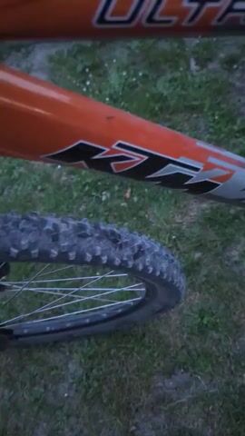 The video shows a close-up view of two bicycles parked on a grassy surface. The bicycles are orange and silver, with visible "ULTRA" and "KTM" markings on the frames. The camera pans across the bikes, focusing on the tires, frames, and handlebars. The lighting suggests it is either dusk or dawn, with the overall scene appearing somewhat dim. No people are visible in the video, and no actions or interactions are depicted.