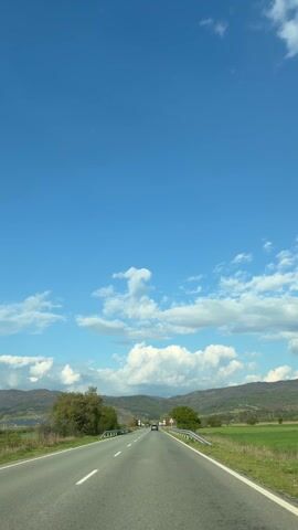 A static, vertical shot captures a road winding towards distant mountains under a bright blue sky dotted with fluffy white clouds. The road is asphalt with white lane markings, bordered by green fields and scattered trees on either side. Guardrails line the edges of the road. Road signs are visible near the middle distance. A single car is approaching in the distance. The overall impression is one of a scenic drive in a rural area on a clear day.