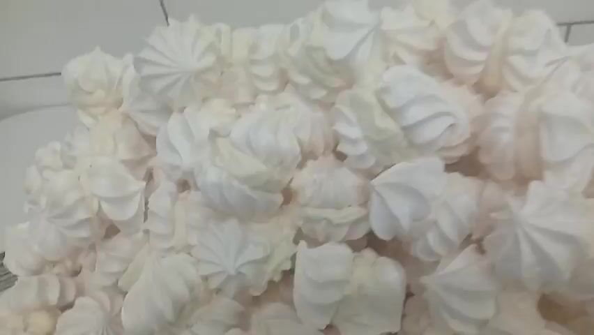 The video displays a close-up, overhead shot of a large pile of white meringues. The meringues are irregularly shaped, with swirled patterns typical of piped meringue. The camera slowly pans to the right, revealing more of the pile. The lighting is bright, and the meringues appear to be on a flat surface, possibly a tray or a countertop. No people or other objects are visible in the frame. The overall impression is of a food production setting, with a large quantity of a sweet treat.